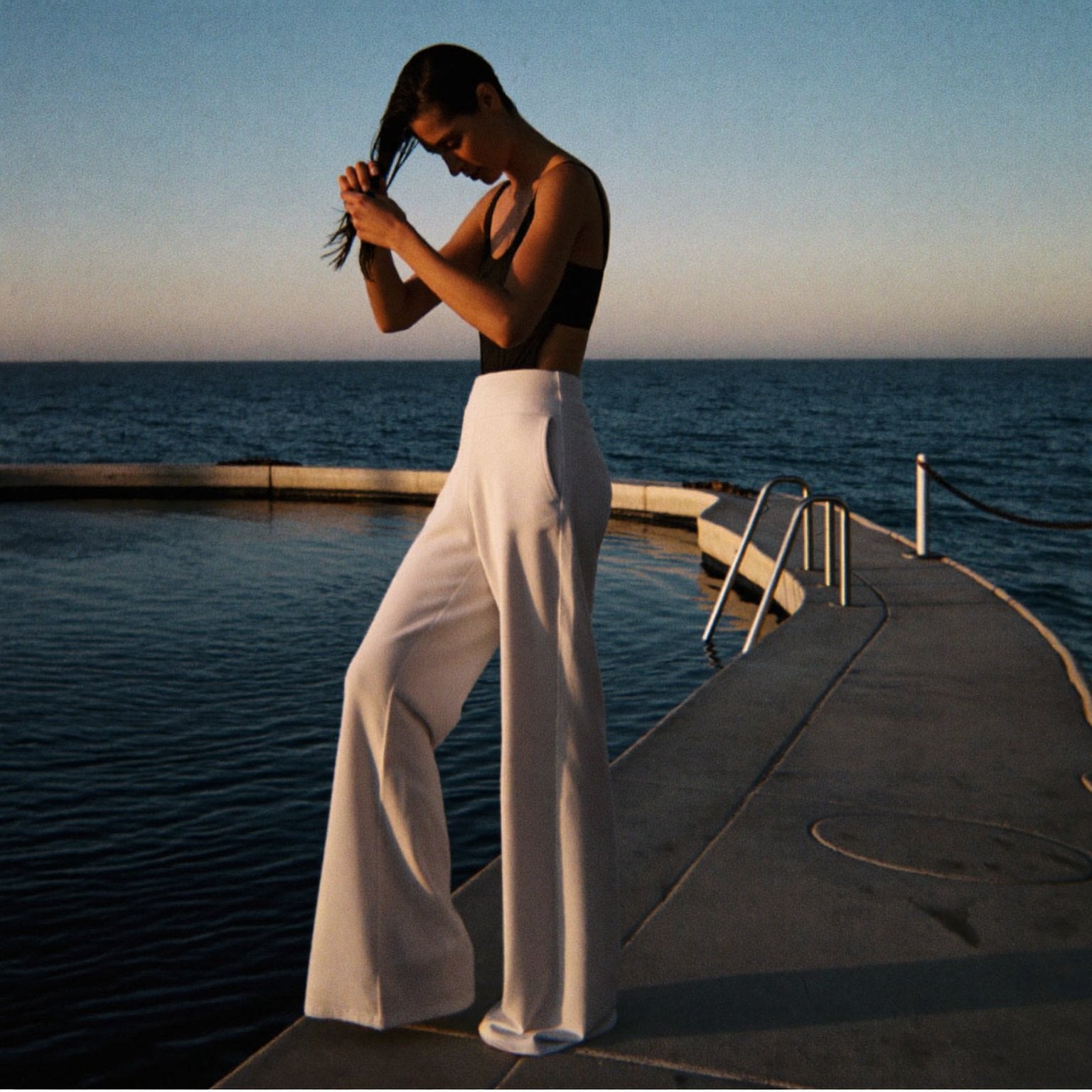 Woman at the edge of Bronte pool at sunrise in white wide-leg pants, wringing out her hair